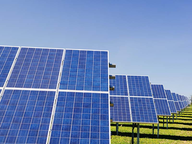 Photorealistic image of a solar panel farm with rows of blue, grid-patterned panels tilted at an angle, mounted on metal poles and anchored in the ground, under a clear blue sky.