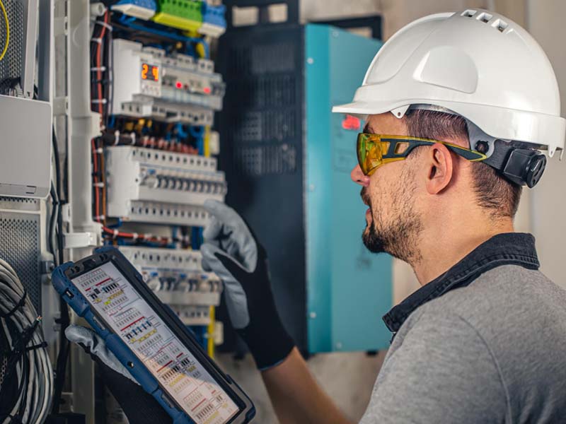 Professional in white hard hat and black gloves using a tablet with a wiring diagram to work on an open electrical panel, revealing the intricate wiring and circuit breakers inside, against a blue wall.