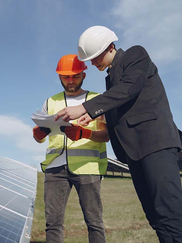 Two professionals, one in a high visibility vest and the other in a suit, both wearing hard hats, discussing plans on a clipboard in a field of solar panels under a blue sky with clouds.