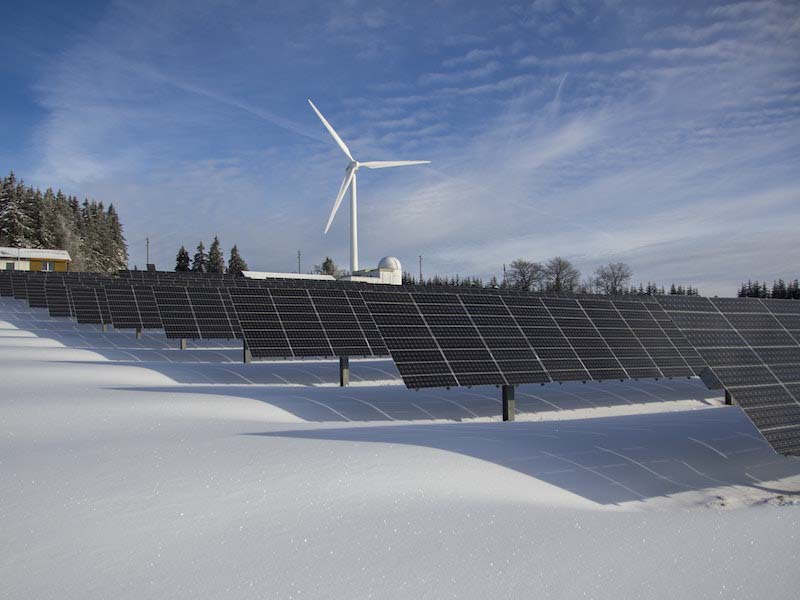 Snow-covered solar panel farm and a wind turbine in a sustainable energy landscape.